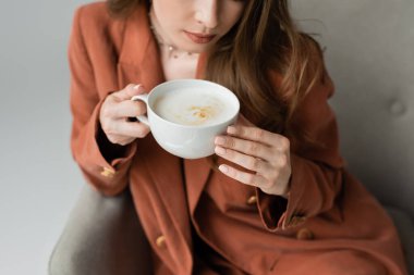 Terracotta blazer ceketli şık, genç esmer bir kadının bulanık ve rahat koltuğunda otururken elinde bir fincan kapuçino tutuşu.  