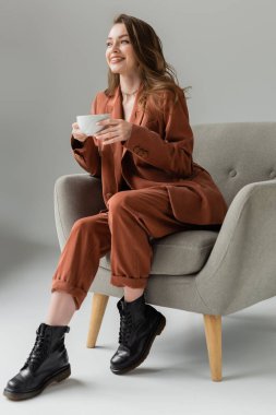 Uzun saçlı ve kolyesi olan mutlu genç bir kadın terracotta giyiyor ve spor ceketli, pantolonlu ve elinde kahve fincanıyla stüdyoda gri arka planda rahat bir koltukta oturuyordu. 