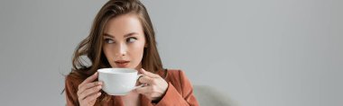 brunette young woman with long hair and necklace wearing trendy suit with blazer and drinking coffee while looking away on grey background in studio, sophisticated lady, coffee break, banner clipart