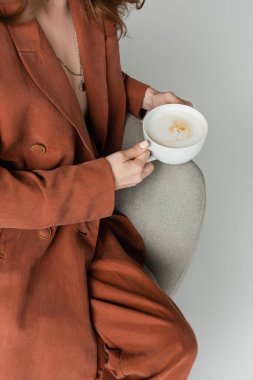 Stüdyoda gri arka planda rahat bir koltukta otururken elinde süt köpüklü bir bardak kapuçino tutan terracotta ve modaya uygun bir takım elbise giymiş genç bir kadın görüntüsü.