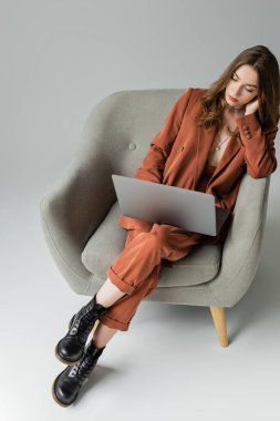 Esmer, uzun saçlı ve kolyeli, spor ceketli ve pantolonlu modaya uygun bir takım giyen gri arka planda koltukta otururken dizüstü bilgisayar kullanan genç bir kadın.