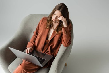Uzun saçlı, terracotta giyimli, spor ceketli ve pantolonlu yorgun genç bir kadın. Gri arka planda rahat koltukta otururken dizüstü bilgisayar kullanıyor. Serbest çalışan, uzaktan kumandalı. 