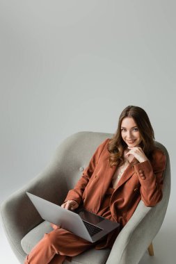 Uzun saçlı ve kolyeli gülümseyen genç kadın spor ceket ve pantolonlu terracotta giyip gri arka planda otururken dizüstü bilgisayar kullanıyor. 