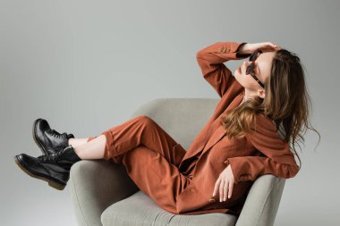 Uzun saçlı şık bir kadın spor ceket, pantolon ve siyah botlarla terasotta takım elbise giyerek gri arka planda koltukta otururken moda olan güneş gözlükleriyle poz veriyor. 