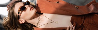 Uzun saçlı, terracotta giyen, spor ceketli ve altın kolyeli modaya uygun giyinen ve soyut gri arka planda güneş gözlükleriyle poz veren güzel model, güneş ışığı, afiş takan genç bir kadın. 