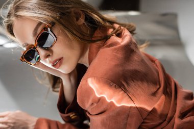 Moda terracotta blazer kumaşında aydınlatma. Uzun saçlı, modaya uygun güneş gözlükleriyle gri soyut arka planda poz veren, güzel model, bulanık arka plan, hayalperest, kameraya bakan genç bir kadın.