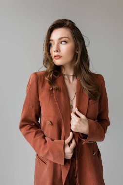 Esmer saçlı, altın kolyeli, terracotta ve şık takım elbiseli, atölyedeki gri arkaplanlı, güzel model, kıvırcık saçlı, modaya uygun saç stili olan şık bir bayan. 
