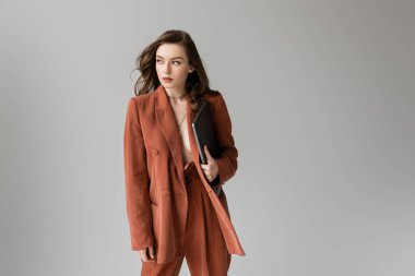 brunette young woman with long hair and necklace wearing terracotta trendy suit with blazer and pants, holding laptop while standing on grey background, freelancer, remote work, looking away clipart