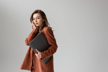 Kumral saçlı ve kolyeli güzel bir kadın terracotta giyiyor ve spor ceketli modaya uygun bir takım elbise giyiyor. Kameraya bakıyor ve dizüstü bilgisayarını tutuyor. Gri arka planda duruyor. 