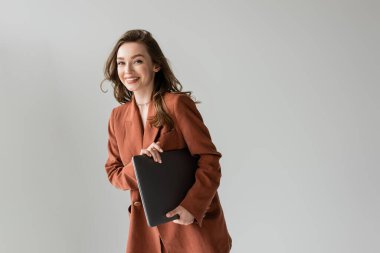 Kumral saçlı ve kolyeli mutlu genç bir kadın kahverengi ve modaya uygun spor ceketli, kameraya bakan ve dizüstü bilgisayarını tutan gri arka planda duran, serbest çalışan, uzaktan kumandalı 