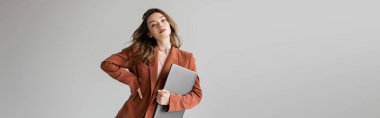 fashionable freelancer with brunette hair in brown and trendy suit with blazer, holding laptop while standing with hand on hip on grey background, looking camera, young woman, remote work, banner  clipart