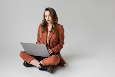 Altın kolyeli çekici bir kadın terracotta ve moda takım elbiseli çapraz bacaklarıyla oturuyor. Gri arka plan üzerinde çalışırken dizüstü bilgisayar kullanıyor. Serbest çalışan, dijital göçebe. 