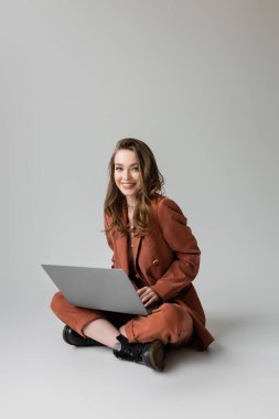 Altın kolyeli mutlu genç bir kadın kahverengi ve moda takım elbiseli çapraz bacaklarıyla oturuyor, gri arka plan üzerinde çalışırken dizüstü bilgisayar kullanıyor, serbest çalışan, dijital göçebe 