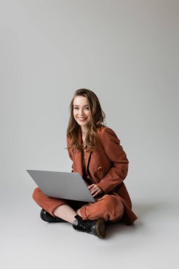 happy young woman in golden necklace sitting with crossed legs in brown and trendy suit, using laptop while working remotely on grey background, freelancer, digital nomad  clipart