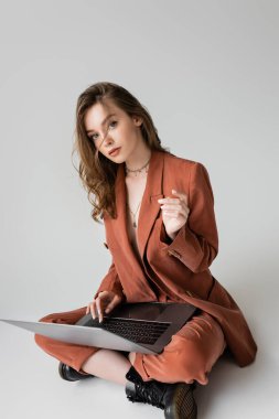 Altın kolyeli, kahverengi ve modaya uygun takım elbiseli genç bir kadın gri arka plan üzerinde çalışırken dizüstü bilgisayar kullanıyor, serbest çalışan, dijital göçebe, kameraya bakıyor.
