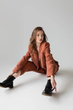 Uzun boylu, esmer, dalgalı saçlı, modaya uygun ve büyük beden takım elbiseli, spor ceketli, pantolonlu ve siyah çizmeli, gri arka planlı, şık görünümlü genç bir kadın.  