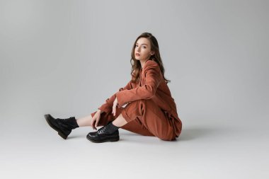 full length of trendy model with brunette and wavy hair sitting in oversize terracotta suit with blazer, pants and black boots, looking at camera on grey background, young woman, stylish pose  clipart