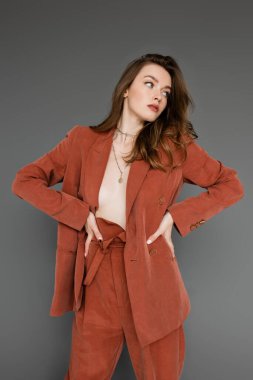 Genç üstsüz, esmer, dalgalı saçlı, elleri modaya uygun ve büyük beden elbiseli, spor ceketli, altın kolyeli, gri arka planlı, şık pozlu, seksi modelli bir kadın. 