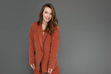 Kumral, dalgalı saçlı, modaya uygun giyinmiş, terracotta blazer ceketli, gri arka planda kameraya bakarken gülümseyen şık bir kadın.