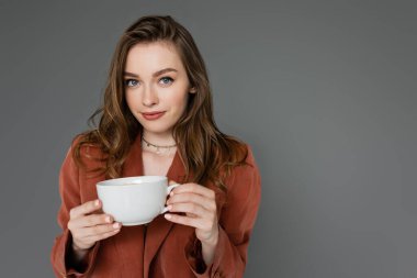 attractive young woman with brunette hair wearing brown and trendy suit with blazer and holding cup of coffee while looking at camera on grey background, work-life balance  clipart