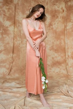 full length of captivating and brunette and young woman in pink silk slip dress holding white flowers while looking at camera while standing on mottled beige background, sensuality, elegance  clipart