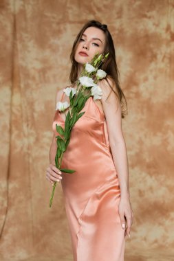 Esmer ve pembe ipek iç çamaşırlı genç bir kadın elinde Eustoma çiçekleri ile dikilip kameraya bakarken benekli bej arka plan, şehvet, zarafet, sofistike 
