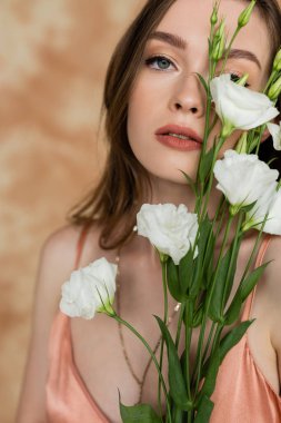 portrait of sensual and young woman holding eustoma flowers while standing and looking at camera on mottled beige background, captivating beauty, elegance, sophistication  clipart