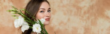 portrait of happy young woman with brunette hair smiling while holding white eustoma flowers on mottled beige background, sensuality, sophistication, elegance, looking at camera, banner  clipart