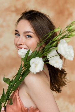 cheerful young woman with brunette hair posing in pink slip dress and holding white eustoma flowers on mottled beige background, sensuality, sophistication, elegance, looking at camera clipart