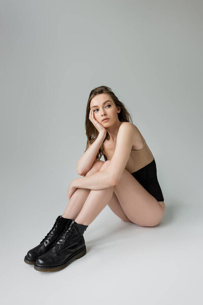 Full length of fashionable young brunette woman in black boots, corset and beige bodysuit looking away while posing and sitting on grey background, dreamy, model
