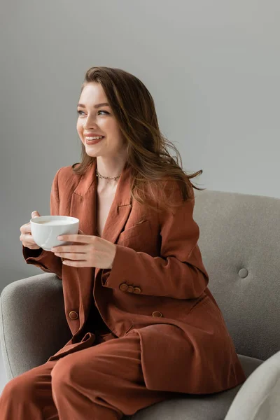 Cheerful young and fashionable woman in terracotta suit and necklace holding cup of cappuccino and looking away while relaxing on modern armchair isolated on grey
