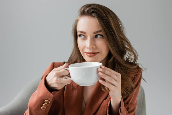 pleased young woman with long hair wearing terracotta and trendy suit with blazer and looking away while holding cup of coffee near blurred comfortable armchair on grey background in studio 