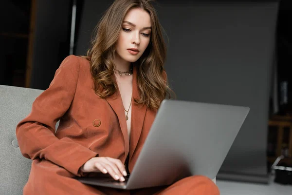 stock image brunette young woman with long hair and necklace wearing terracotta stylish suit with blazer and pants using laptop while sitting in comfortable armchair on grey background, freelancer, remote work 