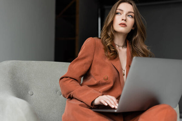 pensive young woman with long hair and necklace wearing trendy suit with blazer and pants and using laptop while sitting in comfortable armchair on grey background, freelancer, remote work 