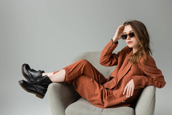 stylish young woman with long hair wearing terracotta suit with blazer, pants and black boots posing in trendy sunglasses while sitting in armchair on grey background, fashionable model, looking at camera