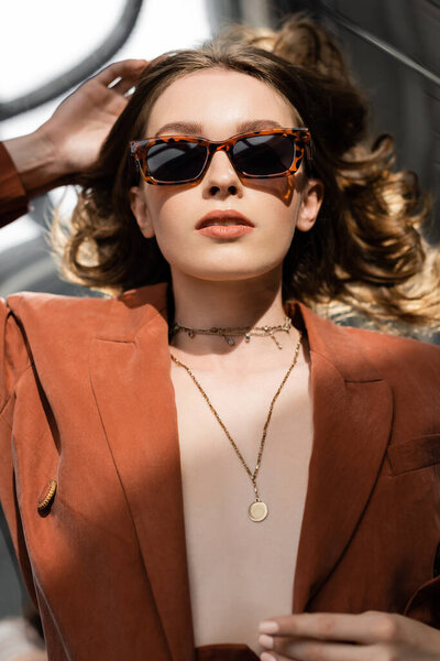 Вид сверху молодой женщины с длинными волосами в терракотовом и трикотажном костюме с блейзером и золотым ожерельем и позирующей в солнцезащитных очках на сером фоне, красивая модель, солнечный свет 