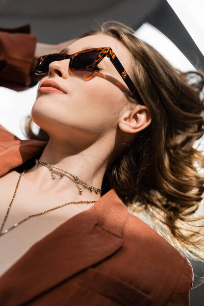 top view of pretty young woman with long hair and natural makeup wearing terracotta and trendy suit with blazer and golden necklace while posing in sunglasses on grey background, beautiful model 
