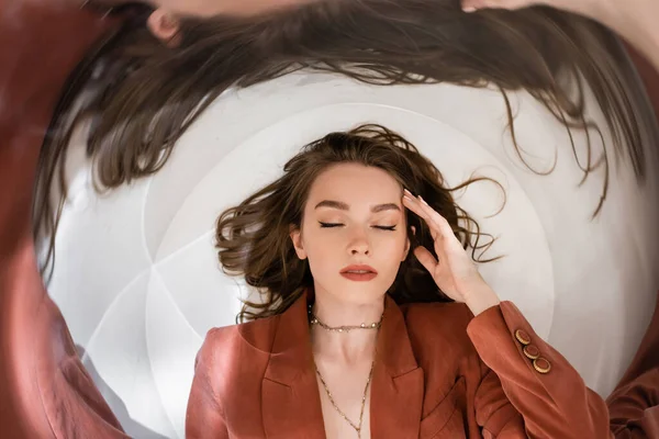 stock image circular shot, top view of young woman with brunette hair wearing blazer and golden necklace while lying and touching face on grey background, blurred, wide-angle, distorted view, closed eyes