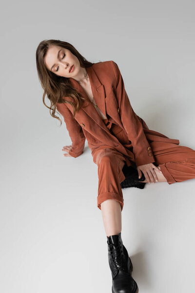 высокий угол обзора модной молодой женщины с брюнеткой и волнистыми волосами, сидящей в модном и крупногабаритном костюме с блейзером, брюками и черными сапогами на сером фоне, стильная модель, закрытые глаза