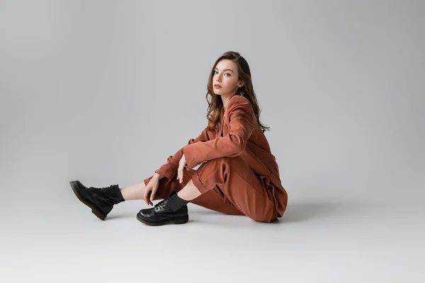 full length of trendy model with brunette and wavy hair sitting in oversize terracotta suit with blazer, pants and black boots, looking at camera on grey background, young woman, stylish pose