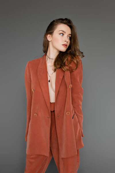young shirtless woman with brunette and wavy hair posing in trendy and terracotta suit with oversize blazer and golden necklace, looking away on grey background,  stylish pose, sexy model 