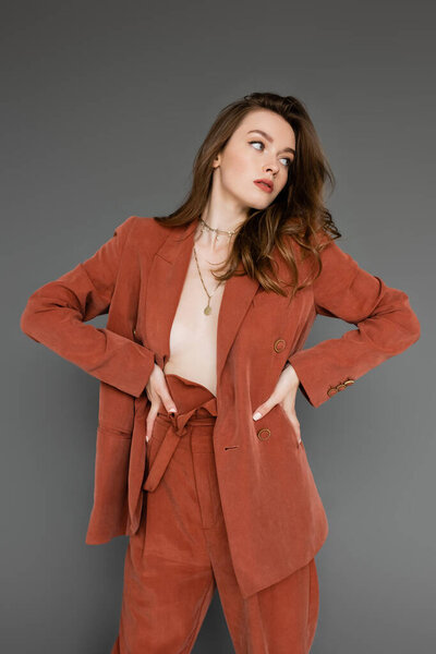 молодая женщина без рубашки с брюнеткой и волнистыми волосами позирует с руками на бедрах в модном и огромном костюме с блейзером и золотым ожерельем, глядя в сторону на сером фоне, стильная поза, сексуальная модель 