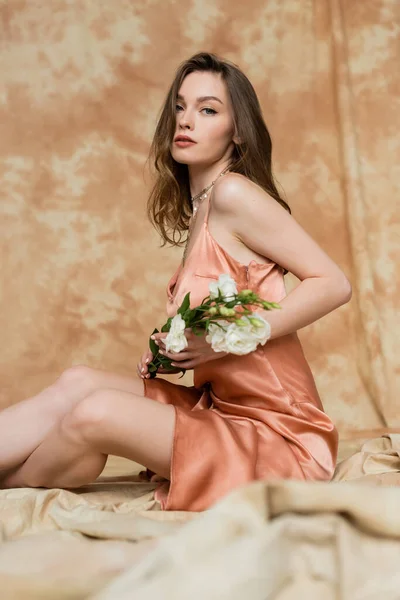 穿着粉色丝滑裙的优雅而漂亮的年轻女子坐在亚麻织物上 拿着点缀过的米色背景 舒适的白花 看着镜头 — 图库照片