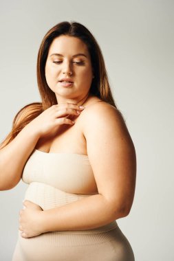 plus size woman with natural makeup posing in beige strapless top and touching shoulder gently in studio isolated on grey background, looking away, body positive, self-love  clipart
