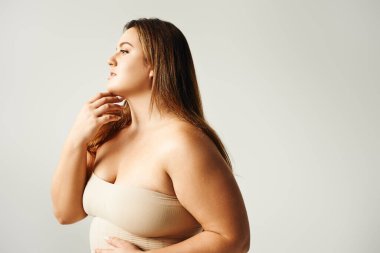 Woman with plus size body touching her chin and looking away while posing in beige strapless top in studio isolated on grey background, body positive, self-love   clipart