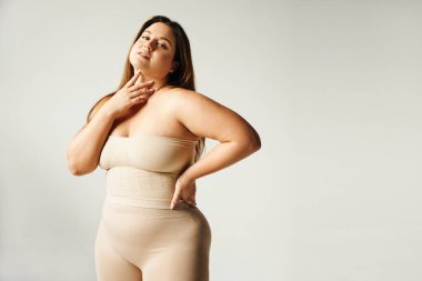 Woman with plus size body touching her neck and looking at camera while posing with hand on hip in beige strapless top and underwear in studio isolated on grey background, body positive, self-love  clipart