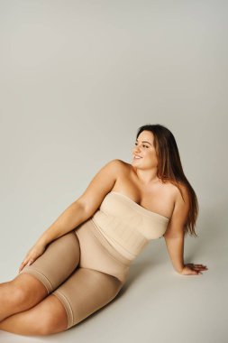 cheerful woman in strapless top with bare shoulders and underwear posing while sitting in studio on grey background, body positive, self-love, plus size, figure type  clipart