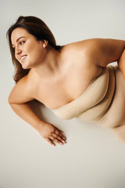 top view of smiling woman in strapless top with bare shoulders and underwear posing in studio on grey background, body positive, self-love, plus size, figure type, looking away  clipart