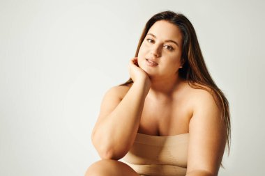 brunette model with plus size body in strapless top with bare shoulders posing while sitting in studio on grey background, body positive, self-love, relaxing, looking at camera  clipart