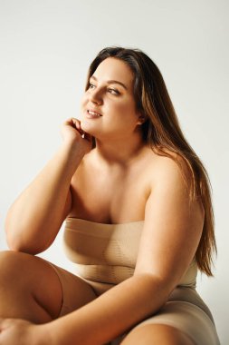 cheerful model with plus size body in strapless top with bare shoulders posing while sitting in studio isolated on grey background, body positive, self-love, relaxing, looking away  clipart